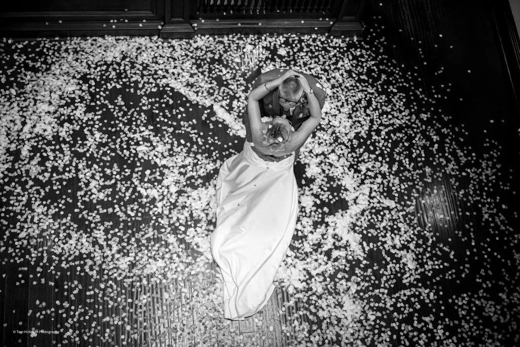 Couple dancing closely on a wooden floor, surrounded by scattered flower petals in a heart shape, with a dark background and floral decor.