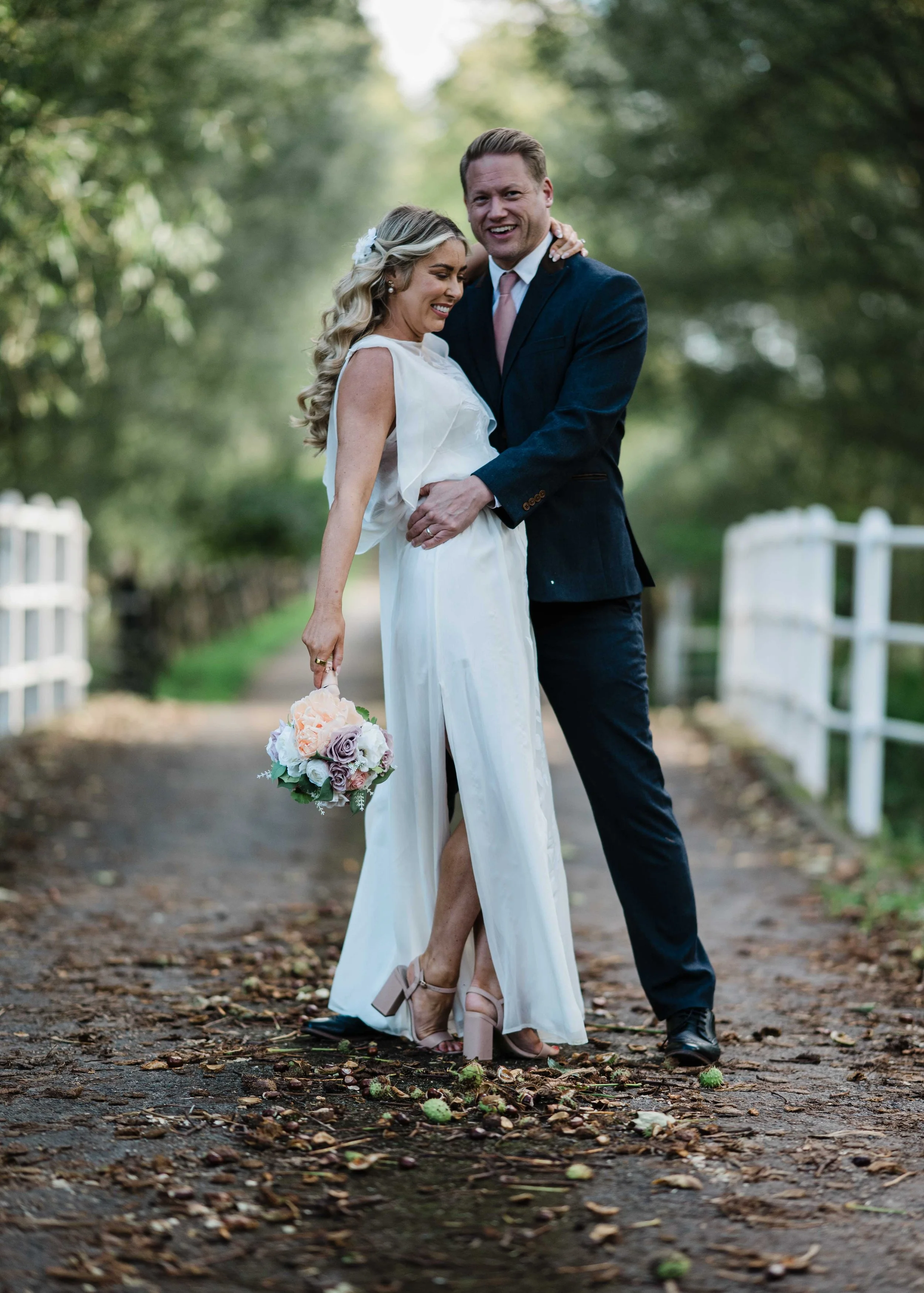 A newlywed couple standing on a wooded pathway during daytime, smiling and embracing each other. The woman is holding a bouquet of flowers and wearing beige high heels, while the man is dressed in a dark suit with a pink tie.