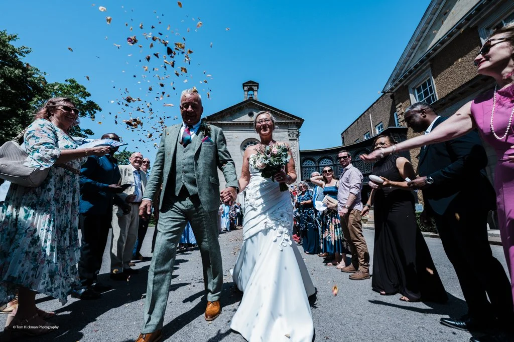 A bride and groom walking hand in hand through friends and family throwing confetti outside a church on a sunny day.