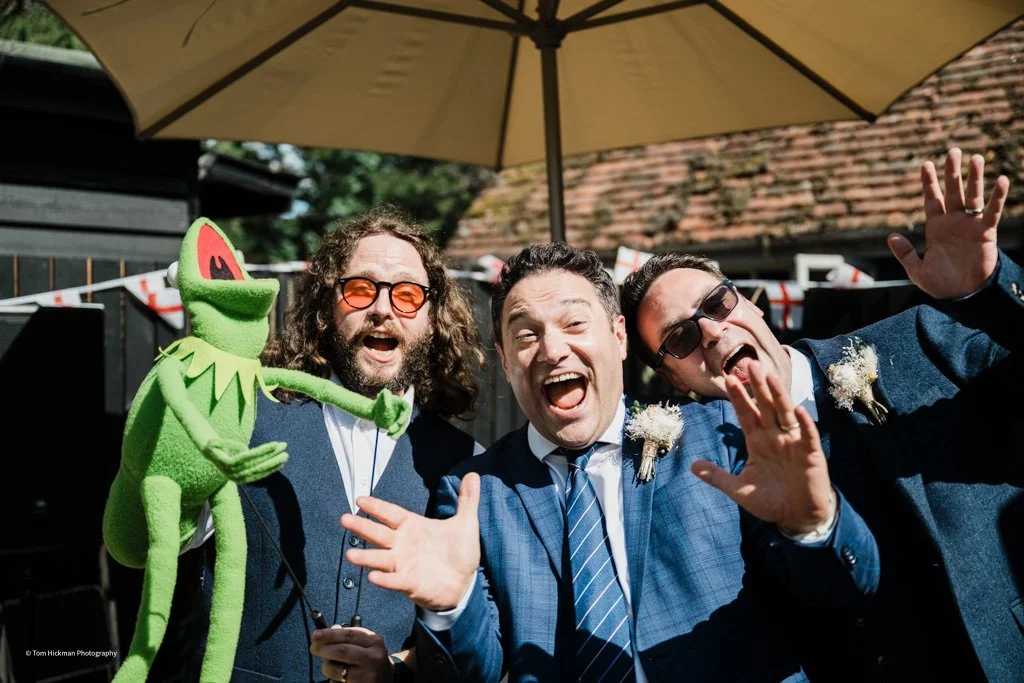 Three men in suits celebrating outdoors under a large umbrella, with a Kermit the Frog puppet, at a festive event with bunting, joyful expressions, and waves.