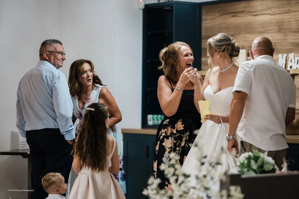 A woman in a floral dress is crying happily while embracing a bride in a white wedding dress. Several other people, including an older man and woman, a young girl, and a young boy, are nearby, watching the emotional moment at a wedding reception.