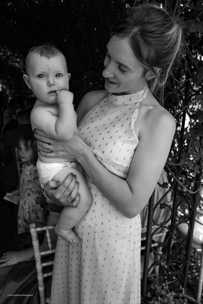 A woman in a sleeveless, patterned dress holding a baby with a cloth diaper, outdoors near a decorative iron fence with foliage. The woman smiles at the baby, who looks at the camera with a finger in their mouth.
