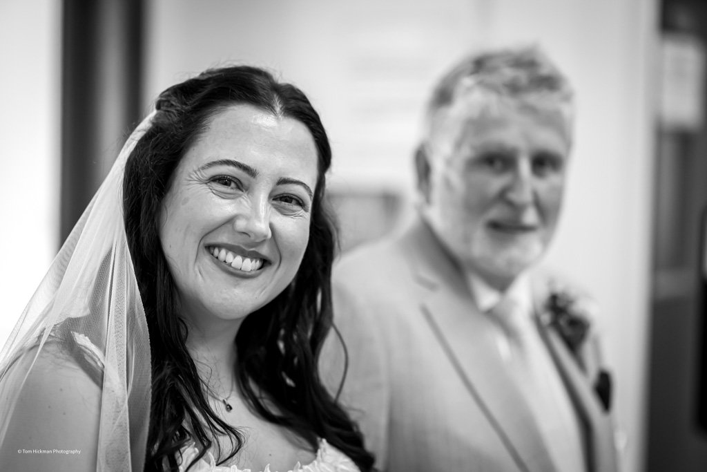 A smiling bride with long dark hair wearing a veil, and a man with gray hair and a beard in a suit, both indoors.