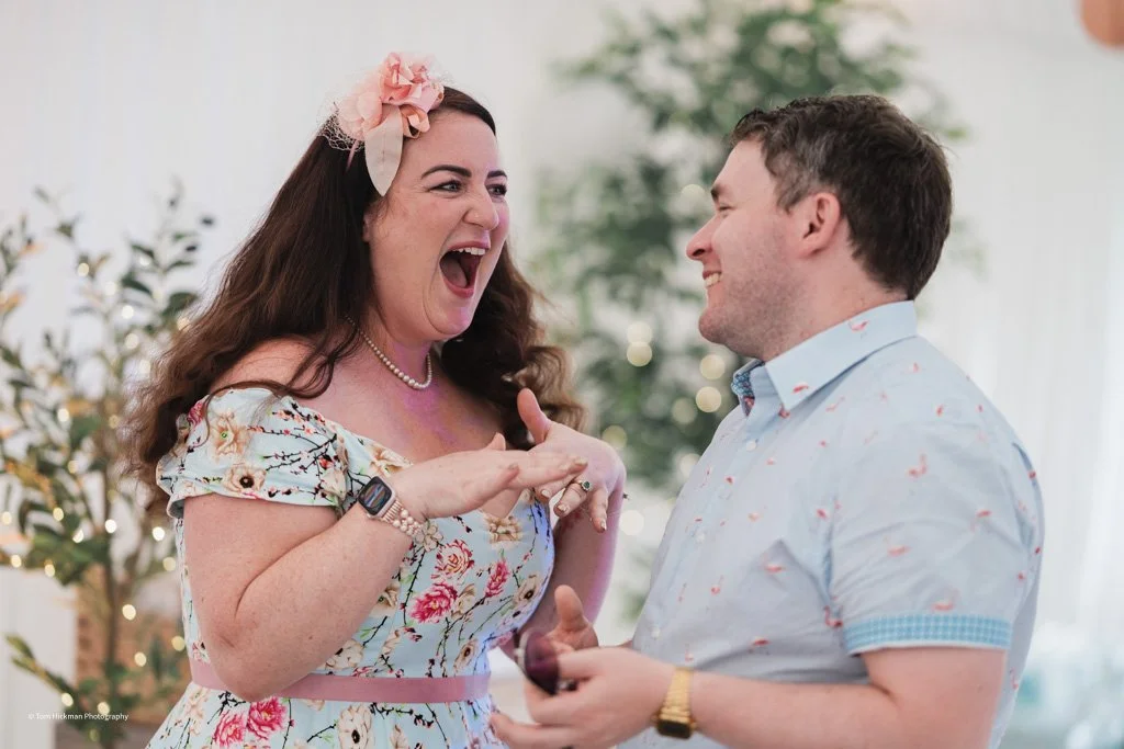 A woman with long brown hair and a pink floral headband laughing and smiling while talking to a man with short brown hair, wearing a light blue shirt, in a decorated room with greenery in the background.