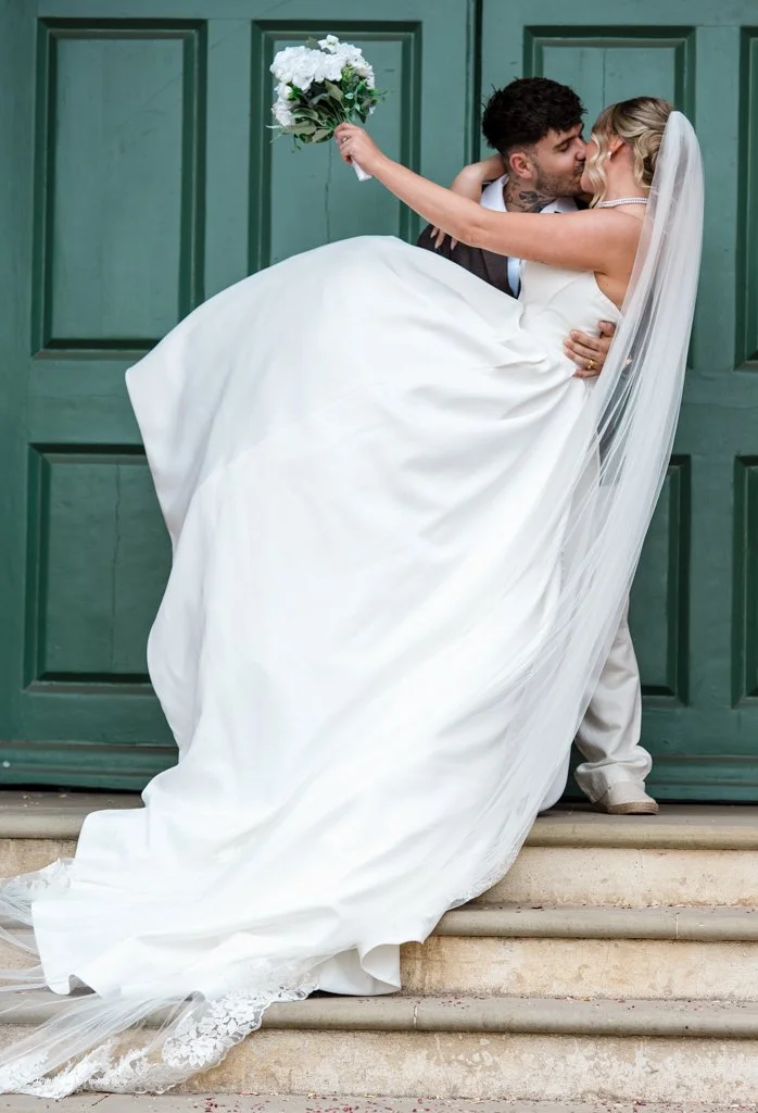 Bride and groom kissing outside green door, bride in white wedding gown holding a bouquet, groom in suit, bride lifting her gown, wedding veil flowing.
