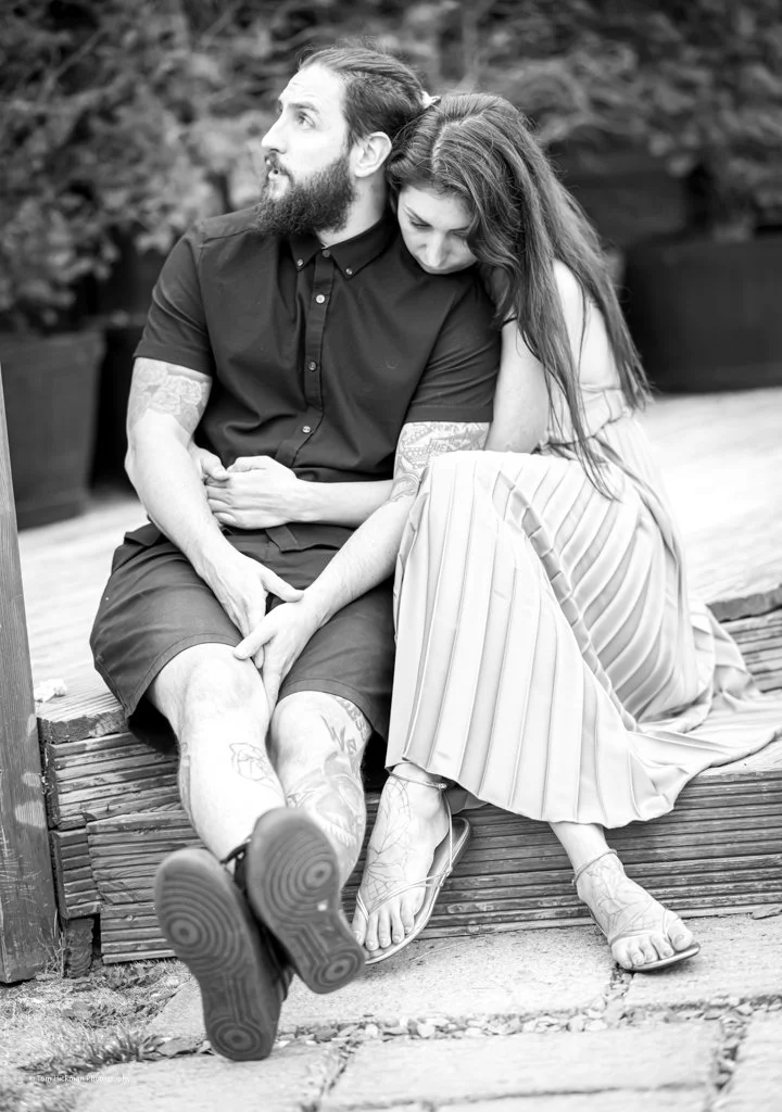 A black and white photo of a man and woman sitting close on a bench, holding hands. The woman rests her head on the man's shoulder and appears to be leaning into him.