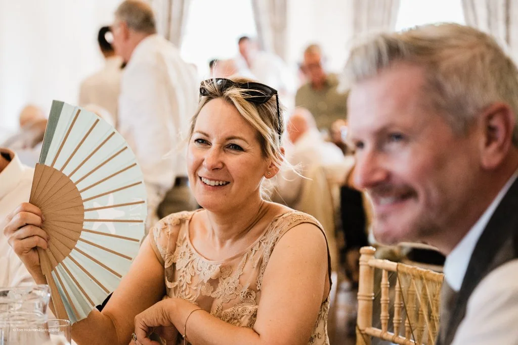 A woman smiling and holding a folding fan at a social gathering or celebration, with a blurred background of other people.