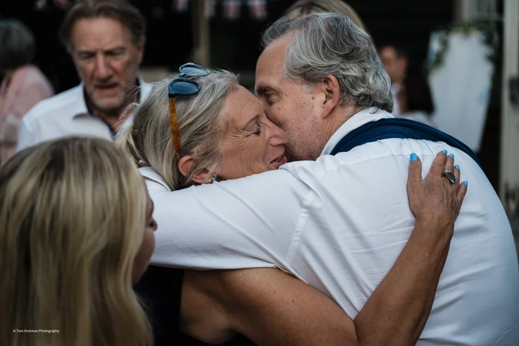 An elderly woman and an older man are hugging each other warmly, smiling as they embrace. Several other people are in the background, appearing to be at a social gathering.