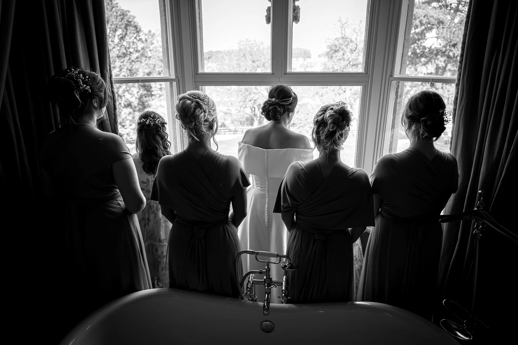 A woman and five girls standing by a large window, gazing outside, with a bathtub in the foreground and curtains on both sides, black-and-white photo.