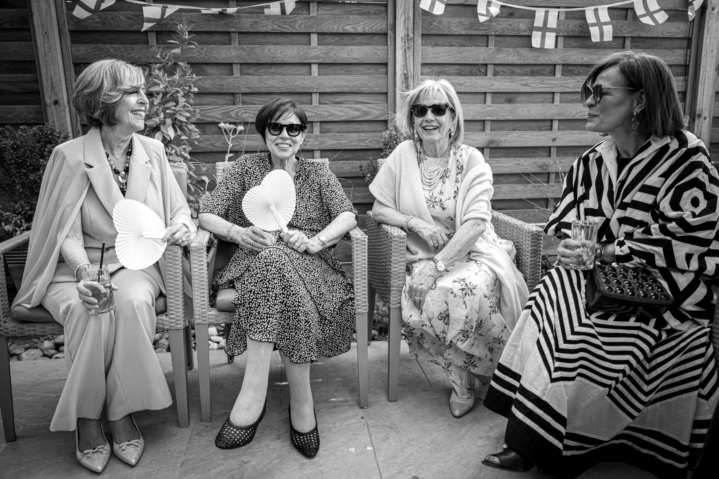 Four women sitting outdoors and engaged in conversation, with a wooden fence and decorative banners in the background, in black-and-white.
