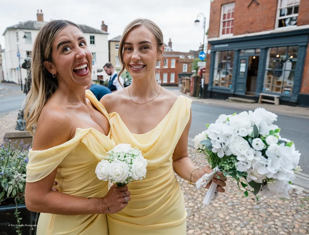 Two women in yellow dresses holding white bouquets standing outdoors on a city street.