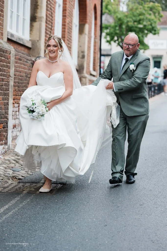 A bride in a white wedding dress and veil holding a bouquet of flowers walks on a city street, assisted by a man in a gray suit with glasses, during a wedding procession.