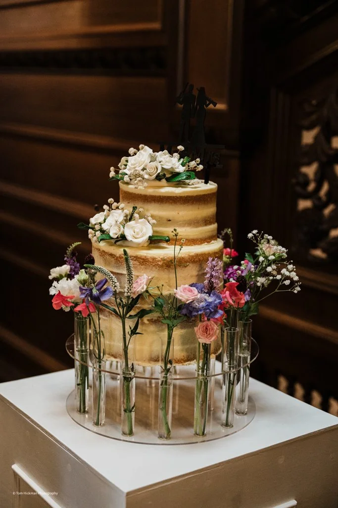A three-tier naked wedding cake decorated with white roses, other small flowers, and greenery, topped with a black wedding cake topper of a bride and groom, surrounded by a circle of small glass vases filled with colorful flowers.