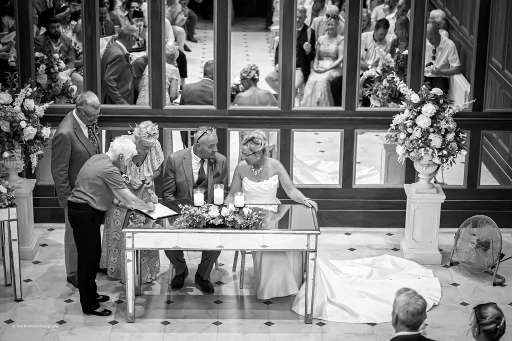 Black and white photo of a wedding ceremony with a bride and groom signing documents at a table, surrounded by family and guests, with floral arrangements and a mirror reflecting additional guests.