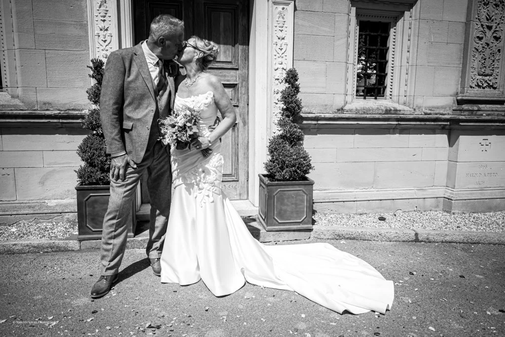 A bride and groom kissing outdoors in front of an ornate building, with potted topiary plants nearby.