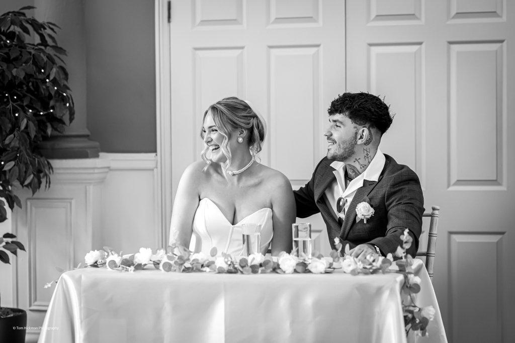 A bride and groom sitting at a table during a wedding reception, smiling and laughing. The bride is wearing a strapless wedding dress and the groom is dressed in a suit with a boutonniere. The table is decorated with flowers and there are bottles of 