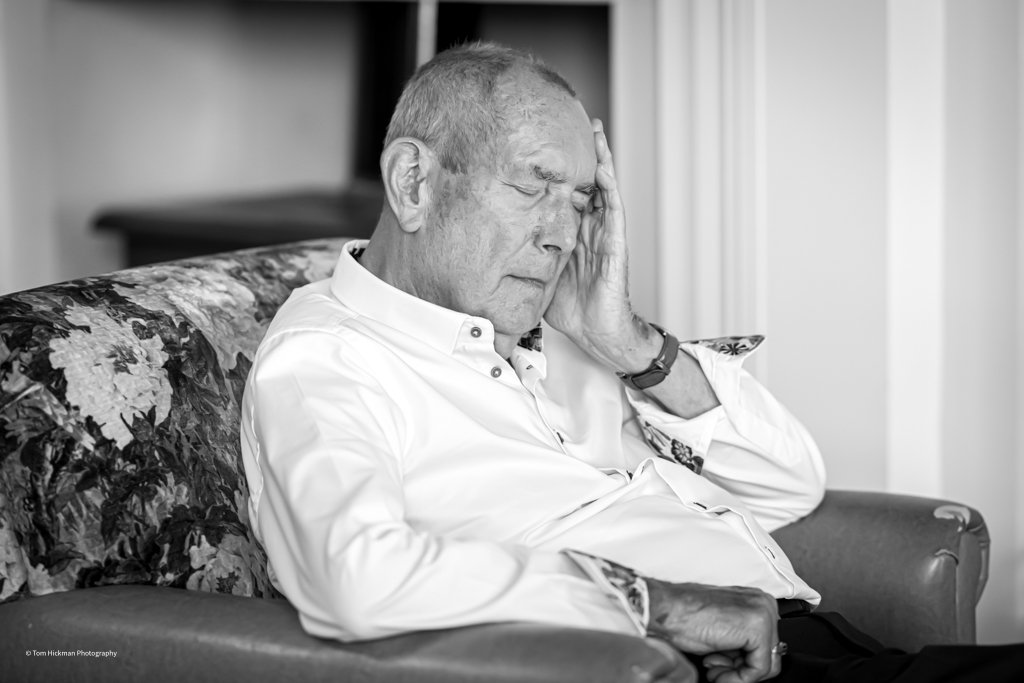 An elderly man with short hair, wearing a white button-up shirt, sits on a patterned sofa with floral upholstery and holds his head with his hand, indicating distress or headache. The photo is in black and white.