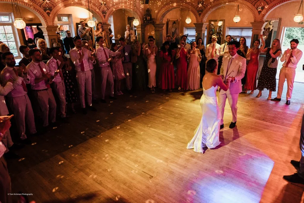 Wedding reception with dancing couple in the center, surrounded by guests clapping and watching in a warmly lit, decorated hall.