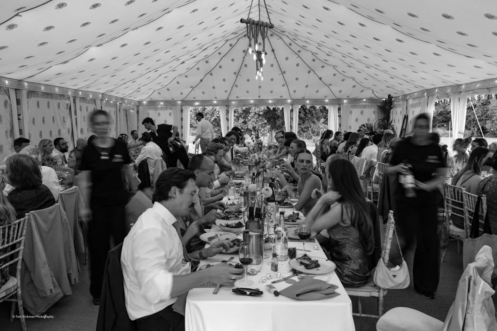 Black and white photo of a large indoor event under a tent, with people seated at long tables, eating, and socializing. Waitstaff are serving and moving around.