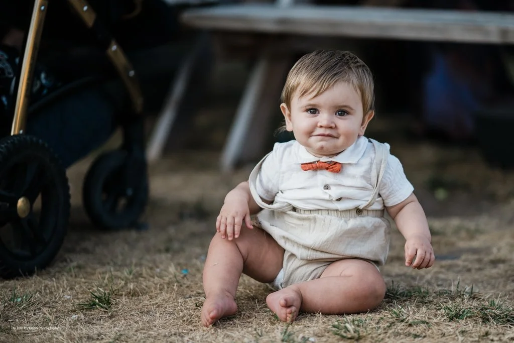 A young toddler sitting on the ground outdoors, wearing a white shirt with a bowtie and beige pants, with a curious expression.