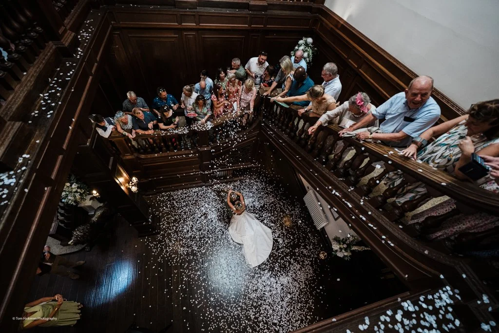 A bride and groom dancing on a dark wooden floor surrounded by guests on a staircase, confetti falling from above.