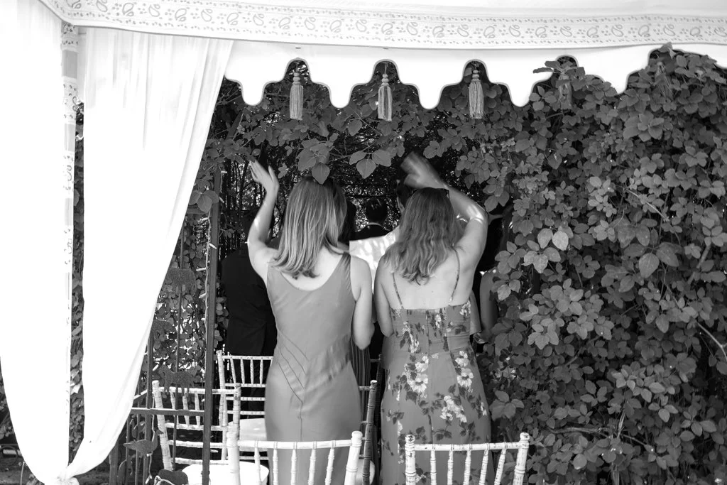 Two women with shoulder-length hair, one in a sleeveless dress and the other in a floral dress, standing under a canopy with curtains, surrounded by leafy bushes, raising their hands.