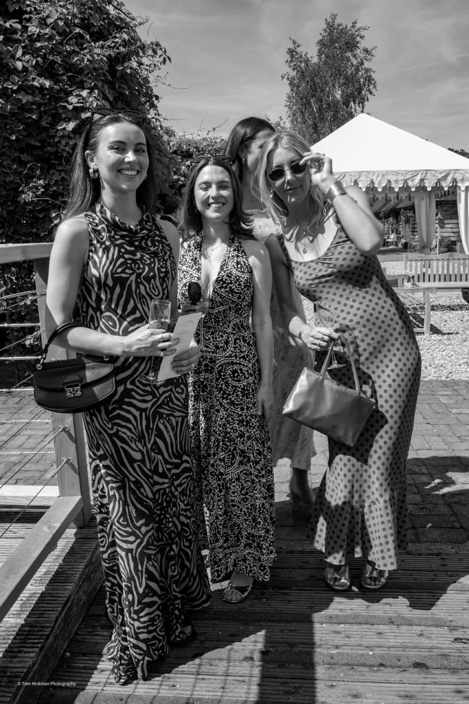 Four women smiling and standing on a wooden deck outdoors, with a tent and trees in the background. They are dressed in summer dresses, some holding drinks and accessories.