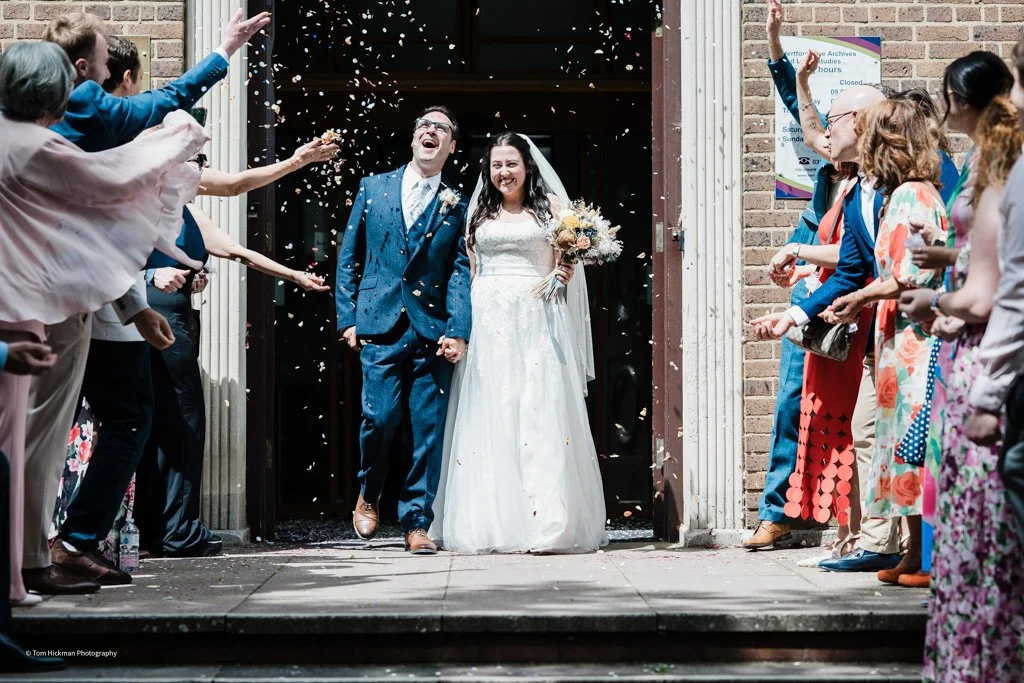Bride and groom exiting a building while guests throw confetti celebrating their wedding.