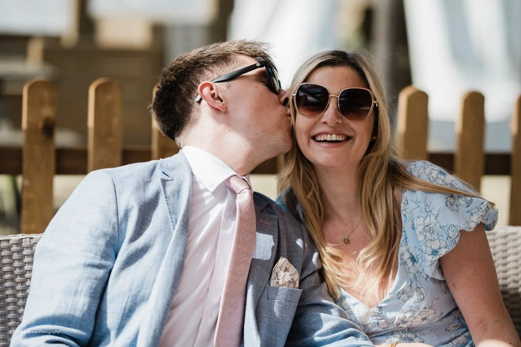 A man wearing sunglasses is kissing a woman with blonde hair and large sunglasses on her face. The woman is smiling and wearing a light blue floral dress.