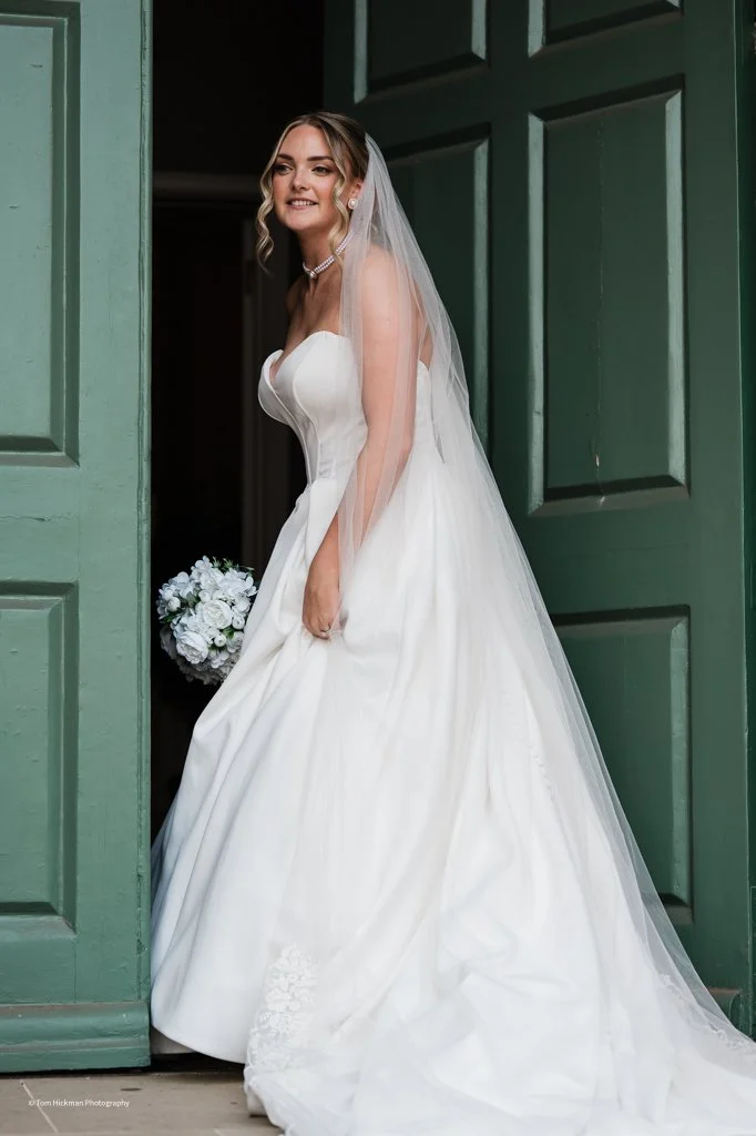 A bride in a white wedding dress with a veil, standing in a doorway, holding a bouquet of white flowers, and smiling.