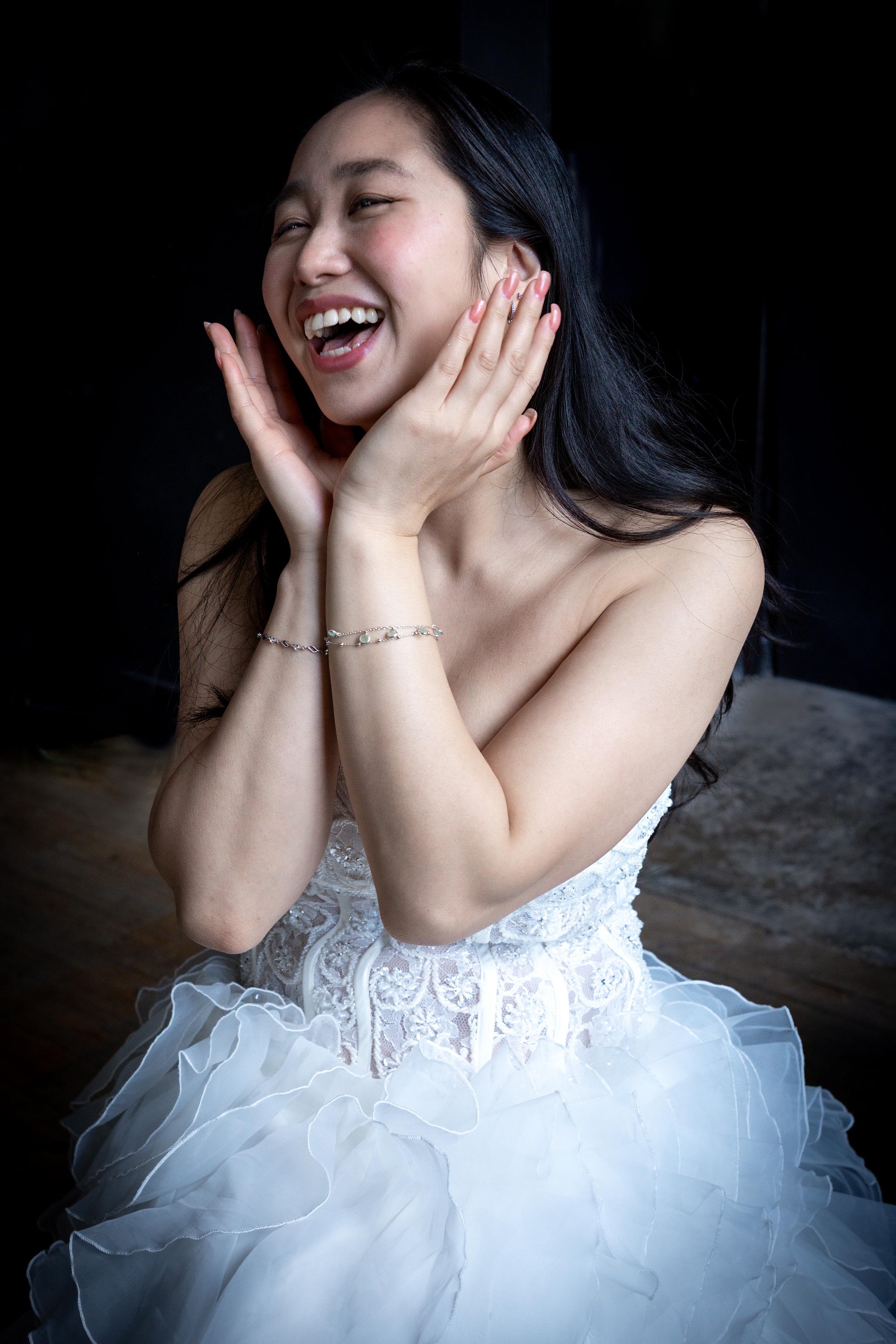 A woman in a white wedding dress smiling and touching her face with both hands, sitting on a dark floor with a dark background.