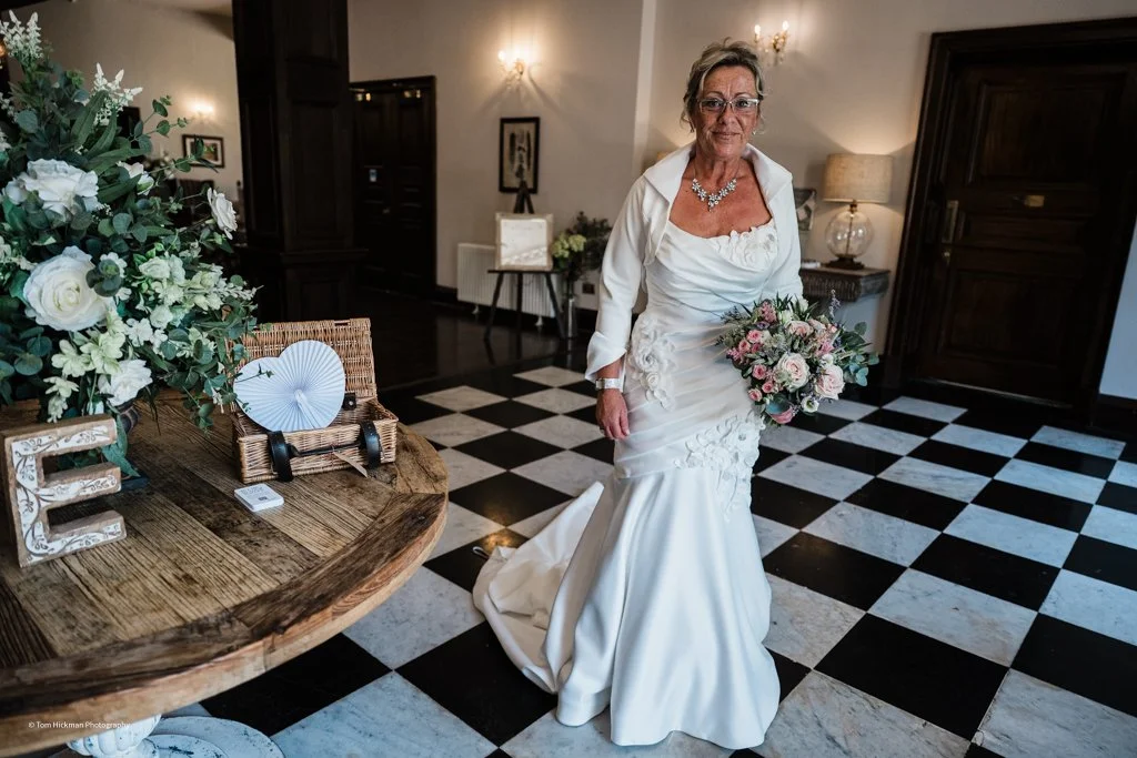 An elderly woman in a white wedding dress holding a bouquet of pink and white flowers, standing indoors with a black and white checkered floor.