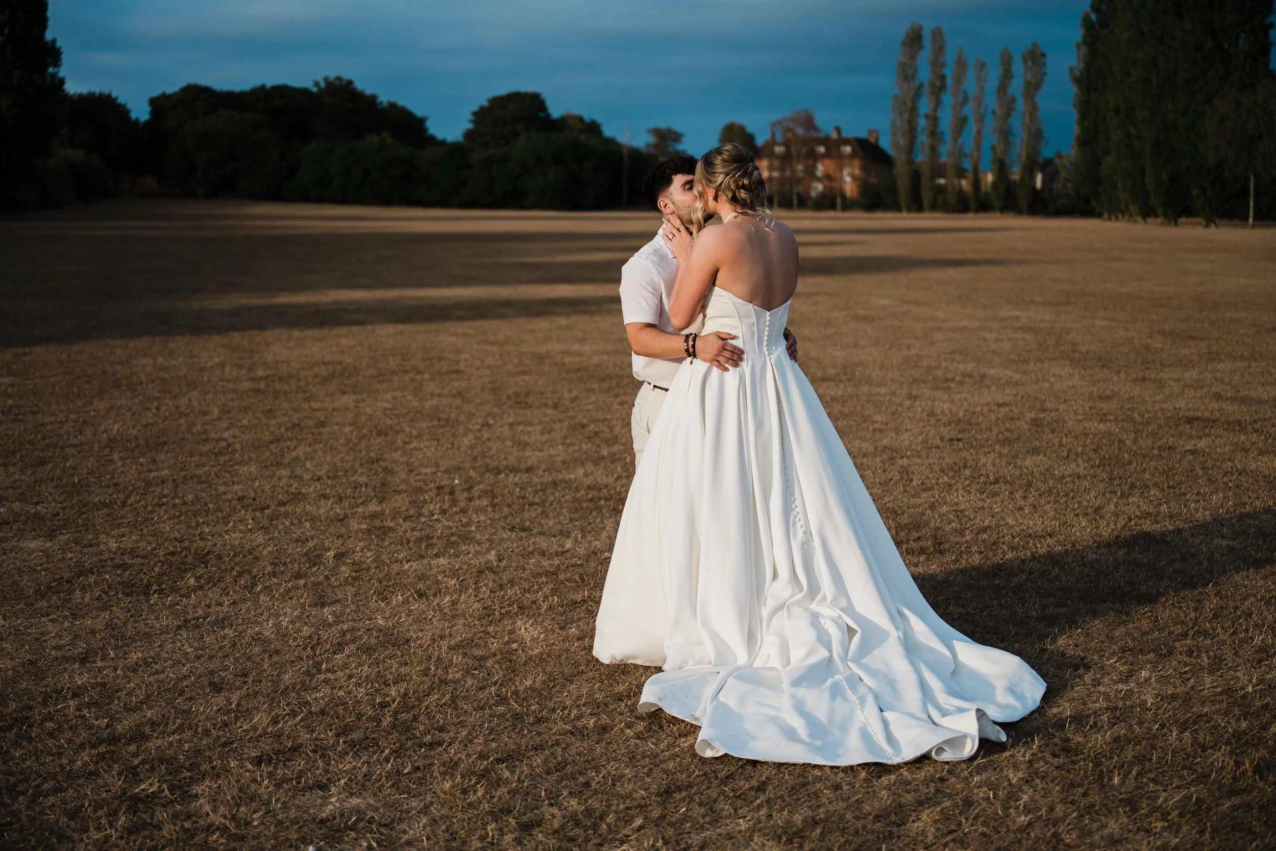 A couple sharing a kiss outdoors at dusk; the woman is wearing a white wedding dress with a long train, and the man is in light-colored clothing. The background shows a field, trees, and a house with a cloudy sky.