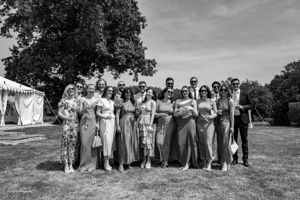 Group of 16 people dressed in formal attire standing outdoors on a grassy field during daytime, smiling for a group photo with tents in the background.