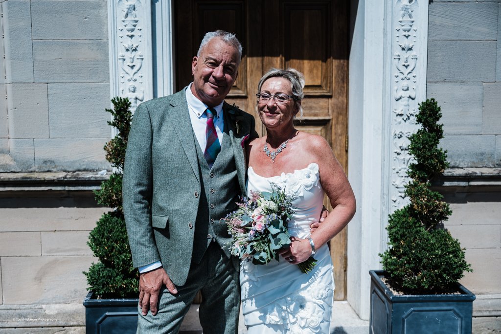 A couple dressed in wedding attire standing outside in front of a wooden door with decorative white trim, with two potted topiary plants on either side.