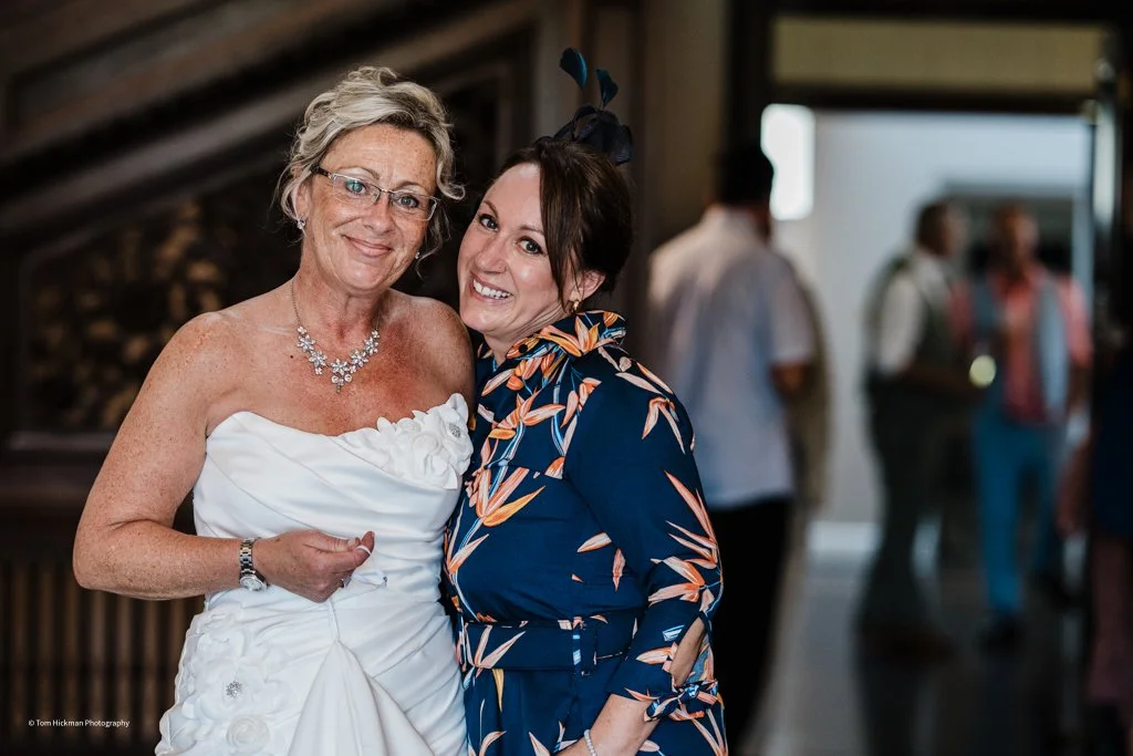 Two women smiling and hugging at a celebration or wedding, one in a white dress with jewelry and glasses, and the other in a floral dress with a hat accessory, with guests in the background.