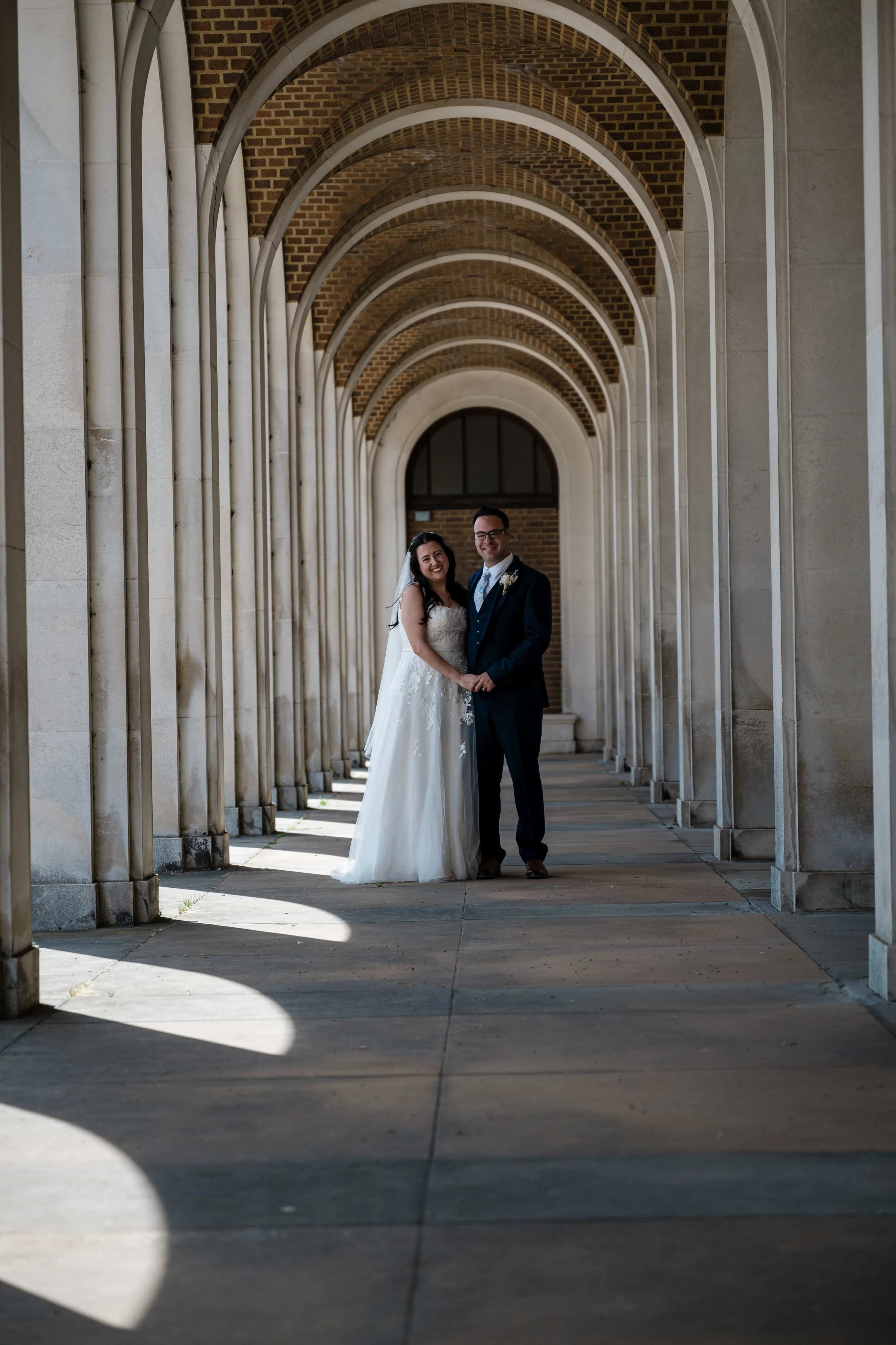 A newlywed couple smiling and holding hands under arched brick and concrete corridor with patterned shadows on the ground.