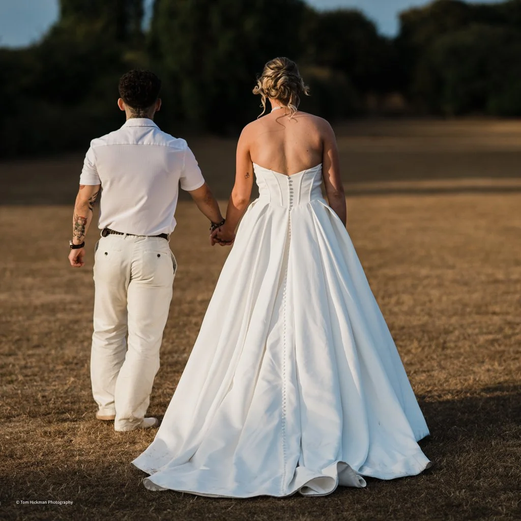 A couple walking hand in hand on a grassy field, with the woman wearing a strapless white wedding gown and the man in a white shirt and light-colored pants.