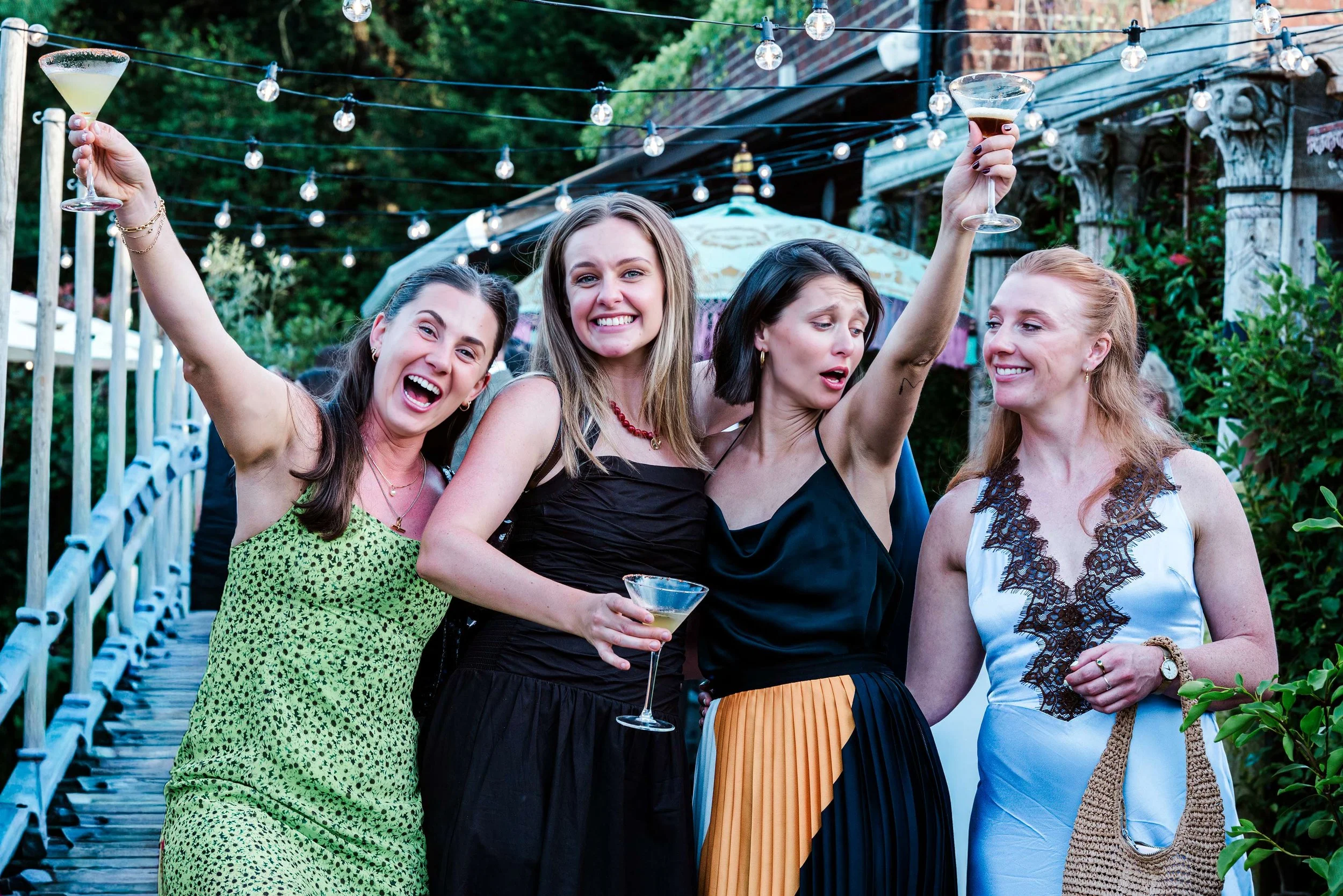 Four women at a backyard gathering, holding drinks and smiling, with string lights overhead and green foliage in the background.