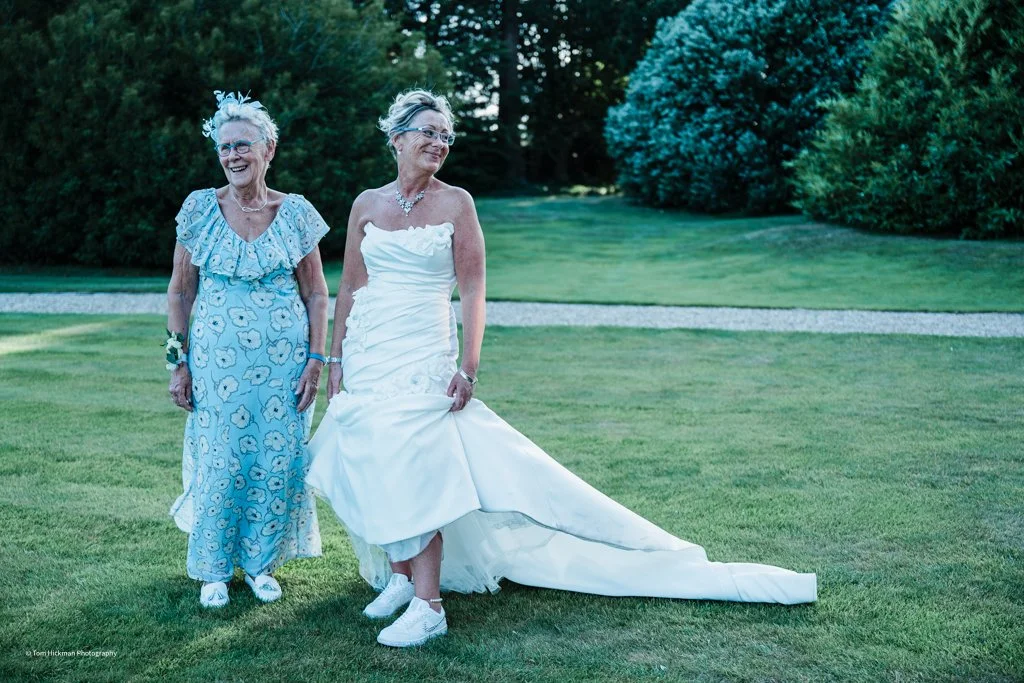 Two women, one in a white wedding dress and the other in a blue floral dress, standing on a lawn with trees in the background during daytime.
