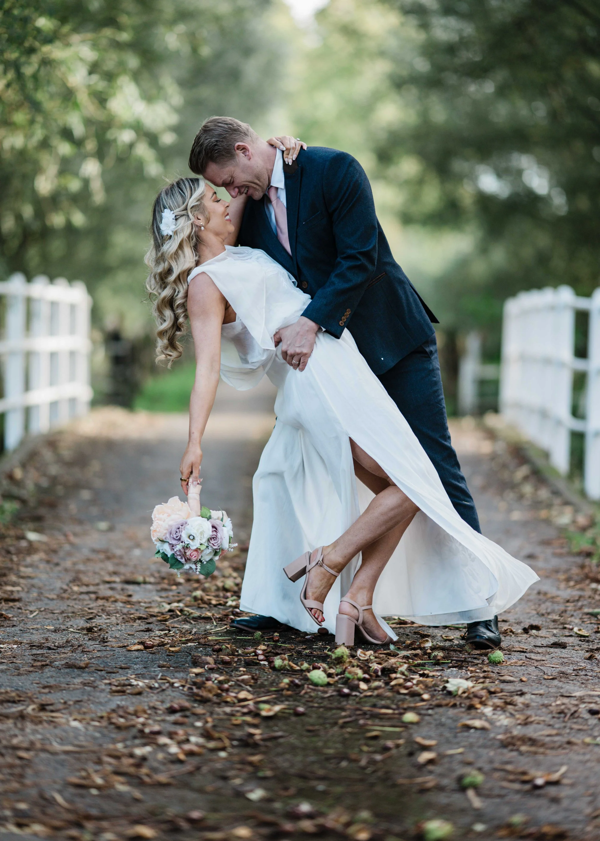 A newlywed couple laughing and embracing outdoors on a tree-lined path with fallen leaves, the groom lifting the bride who is holding a bouquet.