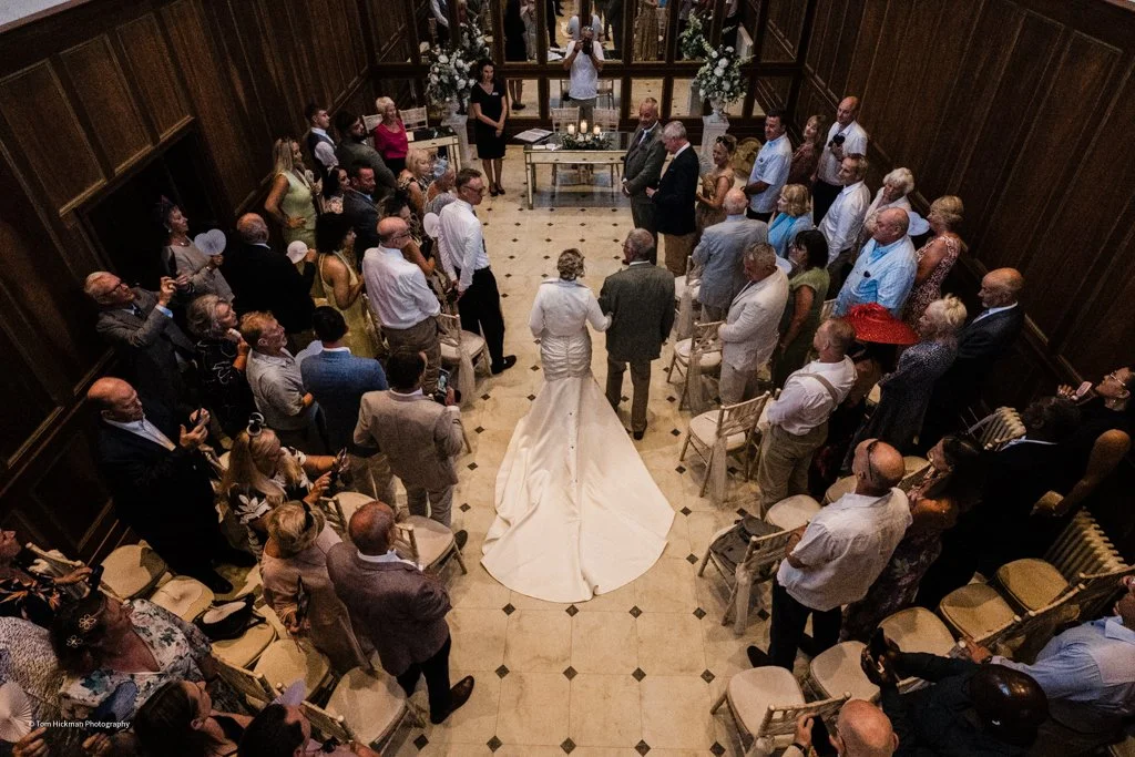 Bride and groom standing in front of officiant, holding hands, in a wedding ceremony with guests seated around them in a wood-paneled room.