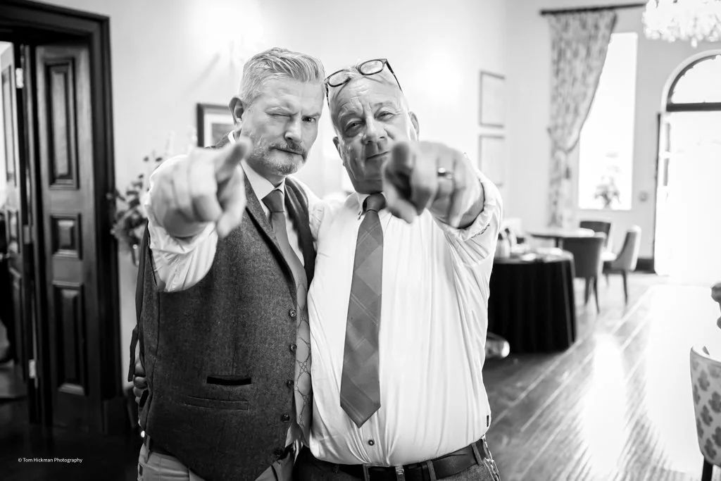 Two middle-aged men in formal attire pointing at the camera in a well-lit room with dining tables and chairs.