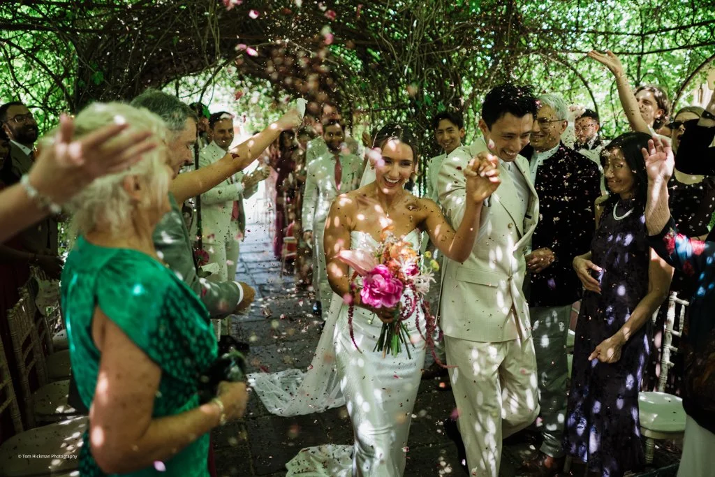 Couple getting married celebrating with guests in a garden, surrounded by greenery, flower petals, and cheerful friends and family.
