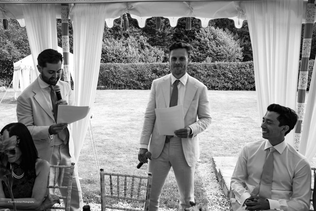 A wedding reception scene with two men in suits, one reading from a paper and the other holding a microphone, under a tent outdoors with greenery in the background. A woman is partially visible sitting nearby.