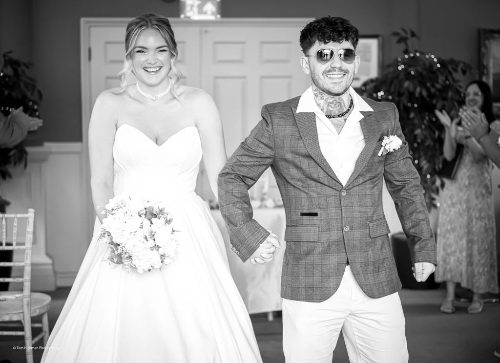 A bride and groom at their wedding reception, smiling and holding hands, with guests clapping in the background.