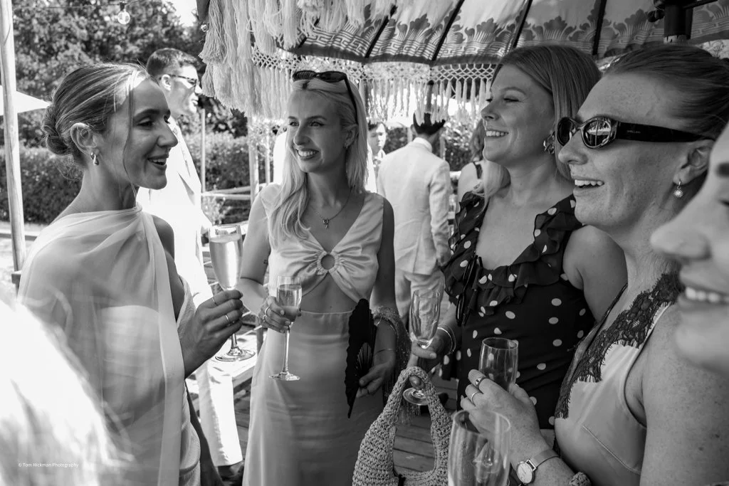 A group of women at a celebration holding glasses of champagne, smiling, and having a conversation outdoors under a canopy.