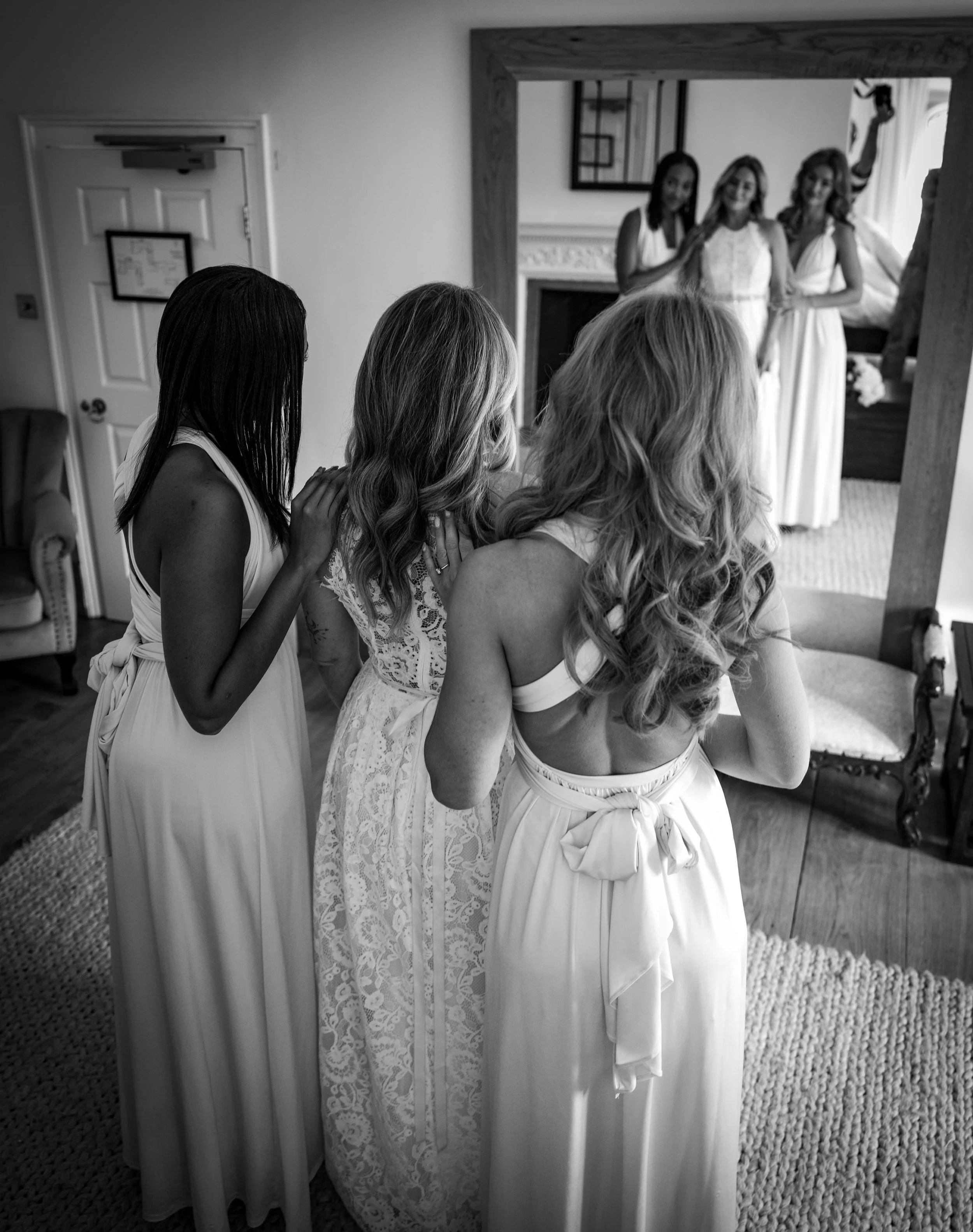 Women dressed in white wedding attire getting ready, taking photos in mirror, in a cozy room with fireplace and decorative mirror.