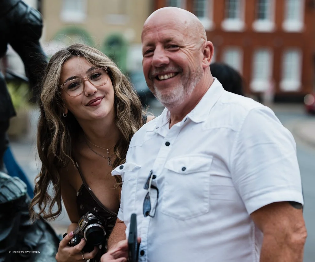 A smiling older man with a shaved head and white shirt standing outdoors, with a young woman with glasses and wavy hair beside him, both in an urban setting.