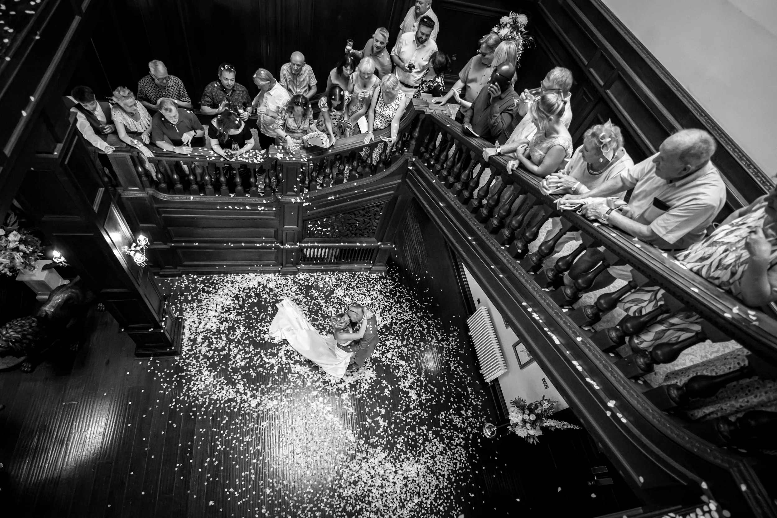 A black and white photo of a wedding dance floor with guests gathered around, watching a bride and groom dance. Flower petals are scattered on the floor, and there are flowers on a table in the background.